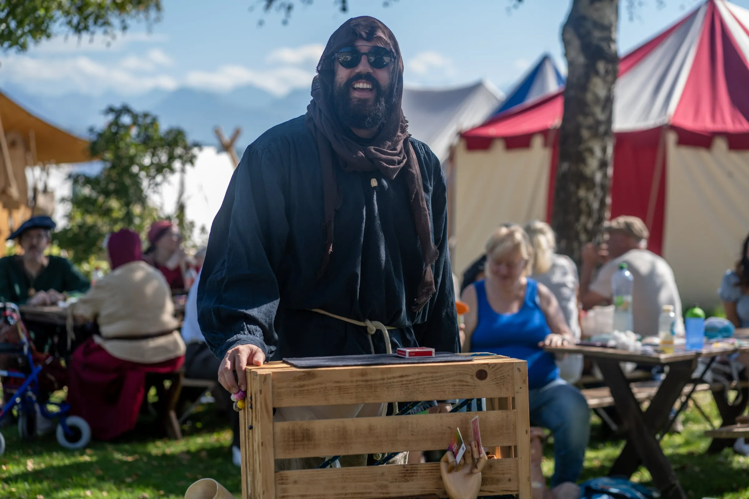 Manuel Kirsch in Gewandung steht an seiner Holzkiste, im Hintergrund Marktbesucher an Holztischen und rot-weisse Zelte.